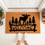 Moose welcome doormat with a rustic forest pine tree design on natural coir fiber, perfect for a cabin porch entry mat or outdoor front door decor, featuring a black silhouette of a moose standing in front of evergreen trees.