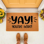 Yay You Are Here doormat featuring bold black text on a natural coir fiber mat, placed on a front porch with yellow flowers and a person wearing brown sneakers standing on the welcome mat for a fun and inviting home entryway