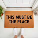 This Must Be The Place coir doormat for front porch entryways, featuring a natural fiber welcome mat with bold black text, perfect for housewarming decor or a stylish home entrance that greets guests with a fun, modern design.