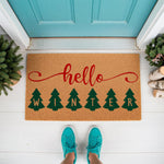 Hello Winter doormat with green pine tree graphics and red script text, featuring a natural coir fiber welcome mat design for seasonal front porch decor that adds a festive touch to your home entrance during the cold weather