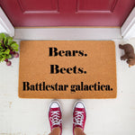 Funny TV show quote doormat featuring Bears, Beets, Battlestar Galactica text on a natural coir fiber welcome mat, perfect for a front porch entry way decor, shown with red sneakers and a small bear figurine on a white painted