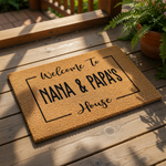 A welcome mat with the text "Welcome To GRANDDAD & GRAMMY'S House" printed on it, surrounded by greenery and a straw hat.