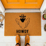 A wooden door mat with a graphic of a cowboy hat and the word "HOWDY" printed on it, surrounded by various floral elements.