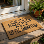 A textured door mat with the text "HOME OF THE WICKED WITCH AND HER LITTLE MONSTERS" printed on it.