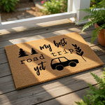 A textured doormat with a black car and tree silhouette printed on it, along with the text "My best road trip yet" and a circular logo with the text "WOOD BY STU - EST. 2010".