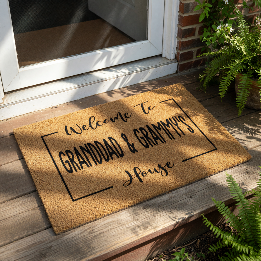 A welcome mat with the text "Welcome To GRANDDAD & GRAMMY'S House" printed on it, surrounded by greenery and a straw hat.
