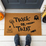 A door mat with the text "TRICK OR TREAT" printed on it, surrounded by various Halloween-themed illustrations such as a ghost, a skeleton, and a candy cane.