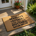 a welcome mat with a humorous message that reads "Welcome to GRANDMA'S KITCHEN * Hope you like * BUTTER & BACON GRAISE!" The mat is placed on a tiled floor in front of a door, with two chairs visible on either side.