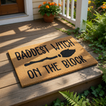 A door mat with the text "Baddest Witch on the Block" printed on it, along with a black silhouette of a broom.
