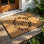A rectangular, beige-colored doormat with a wreath design and the word "Welcome" printed on it.