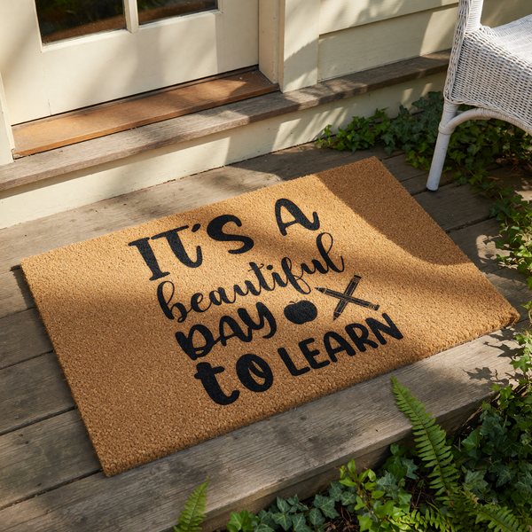 A decorative door mat with the text "IT'S A BEAUTIFUL DAY TO LEARN" printed on it, along with a pair of sandals and a plant.