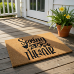 A decorative door mat with the text "Spring is in the air" printed on it, along with various floral and nature-inspired elements.
