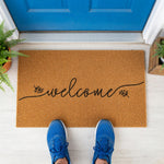 A welcome mat with the word "welcome" written on it, placed on a wooden floor next to a pair of white sneakers.