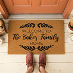 A welcome mat with the text "Welcome to The West Family Home" printed on it, surrounded by decorative leaves and a pair of red sandals.