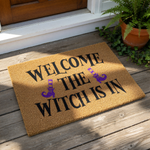 A textured door mat with the text "Welcome The Witch Is In" printed on it, along with two purple witch broomstick icons.