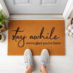 A wooden doormat with the text "Stay while we're Glad You're Here" written on it, placed on a wooden surface.