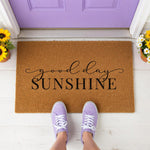 A wooden doormat with the text "good day sunshine" printed on it, placed on a wooden surface with some greenery and red sandals nearby.