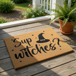 A door mat with the text " Sup witches?" printed on it, along with a silhouette of a witch's hat.