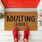 A textured brown doormat with the phrase "ADULTING (KINDA)" printed on it.
