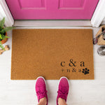A wooden doormat with a decorative design, surrounded by potted plants and flowers on a wooden floor.