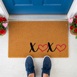 A wooden door mat with the text "XOXO" printed on it, along with two red heart symbols, is placed on a white wooden surface.