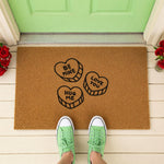 A door mat with a heart-shaped design and the words "Be Mine", "Love Me", and "Hugs Me" printed on it, along with a beaded string and a plant.