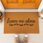 A textured door mat with the phrase "Leave me alone" printed on it, surrounded by a pair of red sandals and some greenery.