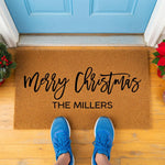 A pair of feet standing on a red and black plaid doormat with the text "Merry Christmas THE MILLERS" printed on it.