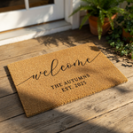 A personalized welcome mat with the text "Welcome The Gussers EST. 2023" printed on it, placed on a brick floor next to a pair of feet and some potted plants.