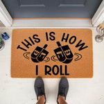 Hanukkah doormat with spinning top design and text This Is How I Roll, a natural coir fiber welcome mat for front porch decor, featuring festive holiday typography and spinning top graphics for a fun seasonal entryway floor rug.