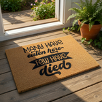 Funny kitchen doormat with the text Many Have Eaten Here Few Have Died printed in black on a natural brown coir fiber welcome mat, perfect for a humorous home decor entry rug or a quirky housewarming gift for a new home owner.