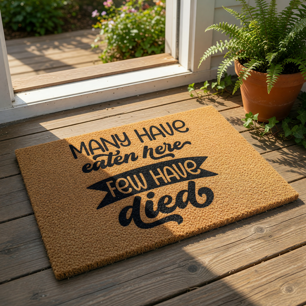 Funny kitchen doormat with the text Many Have Eaten Here Few Have Died printed in black on a natural brown coir fiber welcome mat, perfect for a humorous home decor entry rug or a quirky housewarming gift for a new home owner.