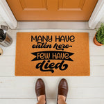 Funny kitchen doormat with the text Many Have Eaten Here Few Have Died, a humorous welcome mat for home decor that features bold black lettering on a natural fiber rug placed on a porch in front of a door with brown shoes visible.