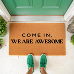 Funny welcome doormat with the text Come In We Are Awesome printed on a natural coir fiber mat, placed at a green front door entrance for a stylish home decor look that serves as a unique housewarming entry rug for your porch.