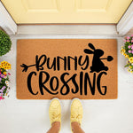 Bunny Crossing coir doormat featuring a black rabbit silhouette and arrow design on a natural fiber welcome mat, perfect for spring porch decor or an outdoor entrance rug placed in front of a yellow door with colorful potted
