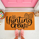 Hunting Crew Easter doormat with black text and egg designs on a natural fiber welcome mat, placed on a porch with a basket of colorful eggs and a bunny figure for a festive spring home decor look at a bright pink front door.