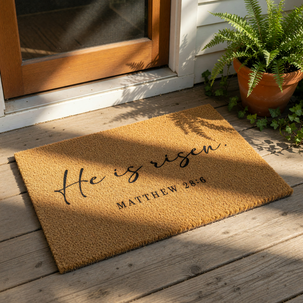 He Is Risen Matthew 28:6 religious doormat featuring natural coir fiber, placed on a porch as a faith-based welcome mat for spring or Easter decor, with script lettering and a clean, durable design for your front door entryway.
