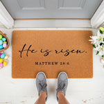 He Is Risen Matthew 28:6 coir doormat for front porch decor, featuring a natural fiber welcome mat with religious scripture text, perfect for spring holiday entryway styling with colorful eggs and white lilies on a white floor.