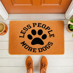 Less People More Dogs doormat featuring a black paw print design on a natural coir fiber welcome mat, perfect for a dog lover front porch decor, placed on a white floor in front of a door with a person wearing orange sneakers