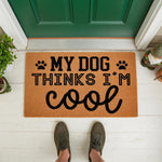 My Dog Thinks I Am Cool doormat, a funny natural coir welcome mat for pet lovers featuring black paw prints and bold typography, placed on a front porch to create a humorous entryway decor piece for dog owners and animal fans.