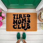 Tired Moms Club doormat featuring bold black retro typography on a natural coir welcome mat, placed on a white porch with green sneakers and flowers, making a funny front door decor piece for any busy mom home entrance.