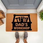 Dad's Workshop doormat featuring a black toolbox graphic with hand tools on a natural coir fiber welcome mat, placed at a front door entrance for a garage or workshop space, making a fun home decor accent for a handy father.