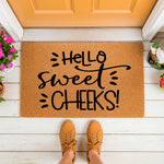 Hello Sweet Cheeks doormat featuring bold black script on a natural coir fiber welcome mat, placed on a white porch floor with a person wearing tan sneakers standing in front of the funny entryway rug for a stylish home decor