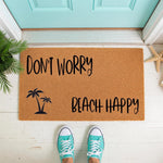 Beach theme doormat with palm tree design and Don't Worry Beach Happy text, a natural coir welcome mat for front porch decor, placed on a white floor with a teal door and starfish accents for a coastal home entryway style.