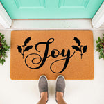 Joy Christmas doormat with holly leaves, a natural coir fiber welcome mat for front porch decor, featuring black script lettering and festive holiday greenery on a tan textured rug surface placed outside a bright teal front door.