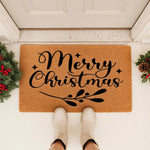Merry Christmas doormat featuring black script text on a natural coir fiber mat, styled on a front porch with festive greenery and pinecones for a holiday welcome mat look that adds seasonal cheer to your home entryway decor.