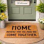 Funny coir doormat with bold black text reading Home Where The Ho And Me Come Together, placed on a round jute rug at a front door entrance, making a humorous welcome mat for a modern home entryway or a cheeky housewarming gift