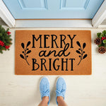 Merry and Bright Christmas doormat featuring festive holiday text and botanical sprigs on a natural coir welcome mat, displayed on a front porch with seasonal greenery and pinecones for a stylish winter home entryway decor look.