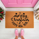Merry Christmas and Happy New Year doormat featuring black script text on a natural coir welcome mat, placed on a porch with pink sneakers, festive greenery, and wood logs for a seasonal holiday home entryway decor look.