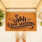Shh Baby Sleeping doormat for front porch, a natural coir welcome mat with black text that alerts visitors to be quiet, perfect for new parents and nursery decor, placed on a doorstep with a person standing in front of the